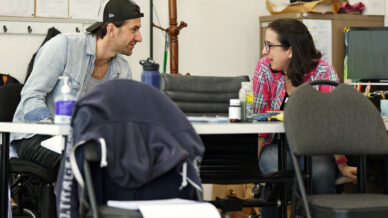 Two people sit at a cluttered table having an engaged conversation in an office or classroom. Both appear relaxed; one wears a backwards cap, the other glasses and a checked shirt. Office chairs and supplies fill the scene.