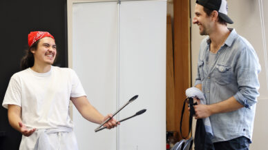 Two people stand indoors, smiling and laughing together. One, wearing a red bandana and apron, gestures with metal tongs, whilst the other, dressed in a denim shirt and cap, holds a towel and looks on.