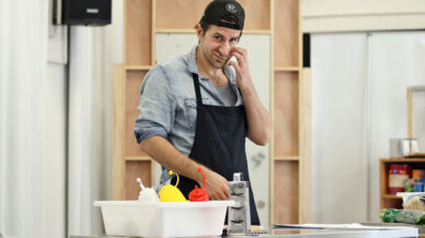 A man wearing a backwards cap, denim shirt, and black apron stands at a kitchen counter with condiments and utensils, touching his ear and smiling, with wooden shelves and a mirror in the background.
