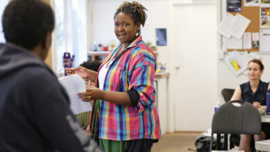 A woman in a colourful checked shirt stands holding papers and addresses a seated group in a casual classroom or office setting. Other people are seated at tables, listening and interacting.