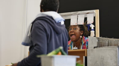 A woman stands behind a counter, speaking animatedly to a person in the foreground. The woman appears expressive, with her mouth open, while food ingredients and order dockets are visible nearby.