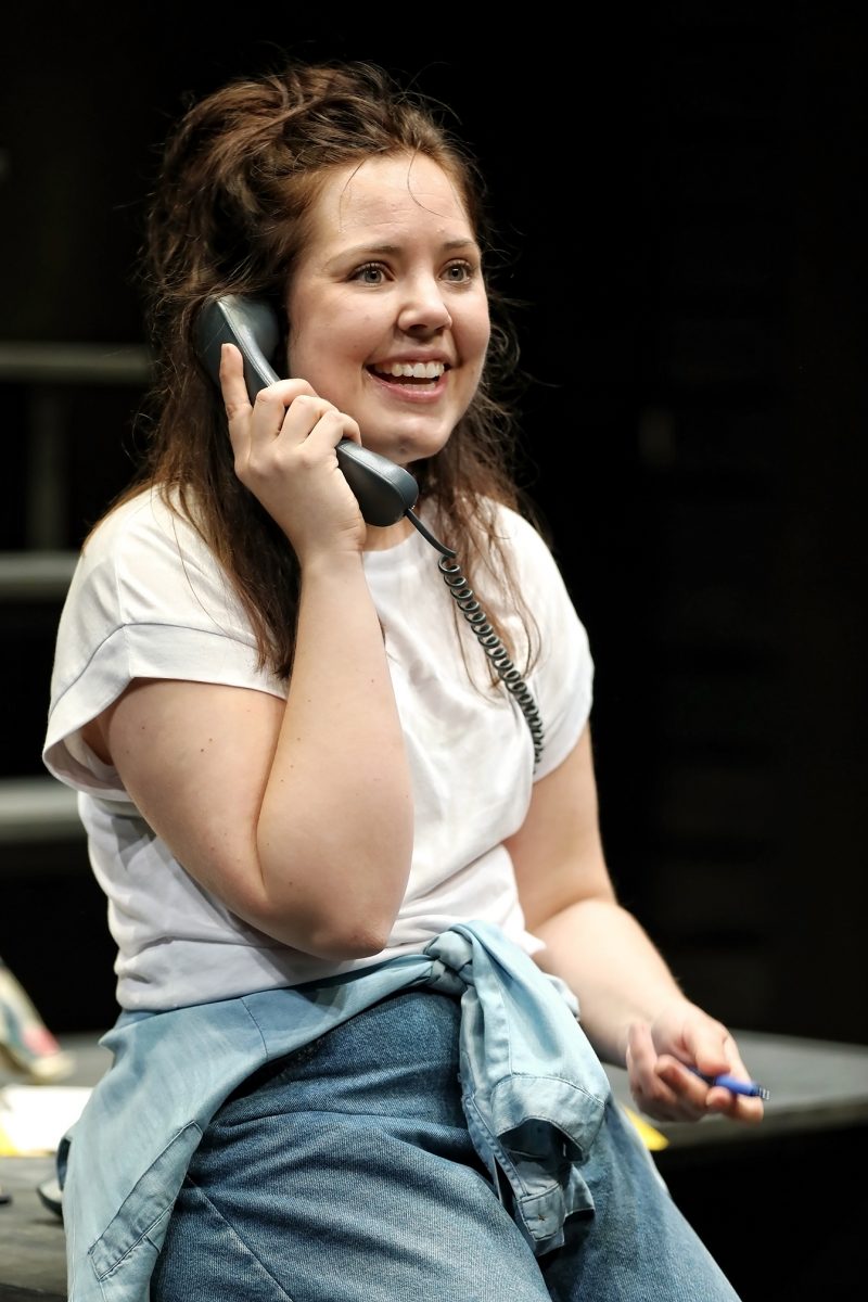 A young woman with wavy brown hair, wearing a white T-shirt and blue jeans with a jacket tied round her waist, smiles whilst talking on a corded phone and holding a pen.
