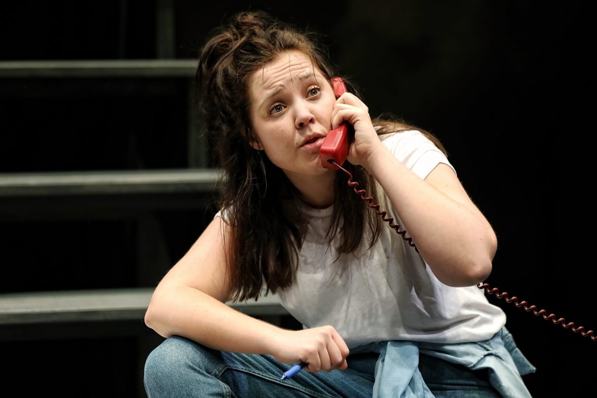 A woman with long brown hair, wearing a white T-shirt and jeans, crouches while speaking on a red corded telephone, looking upwards with a concerned expression. She holds a pen in her other hand.