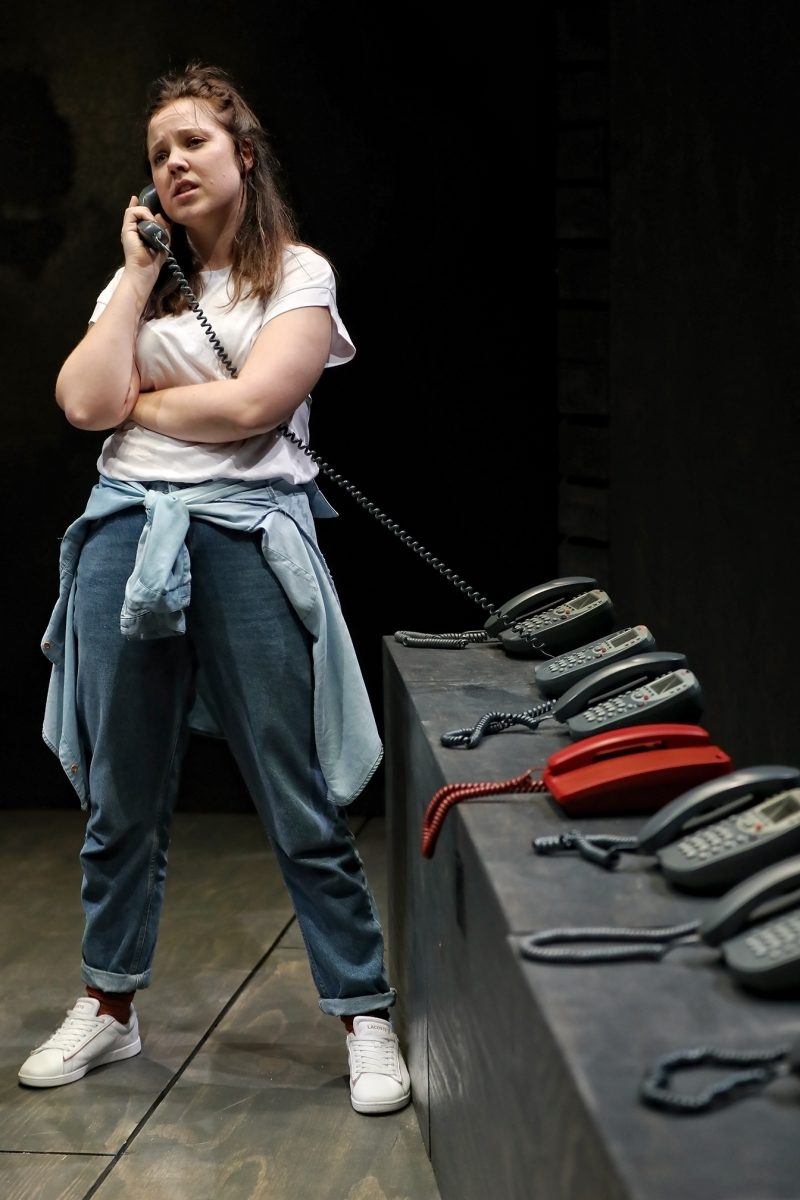 A woman in casual clothes stands by a row of corded telephones, holding one to her ear and looking concerned. Several telephones of different colours are lined up on a dark counter in front of her.
