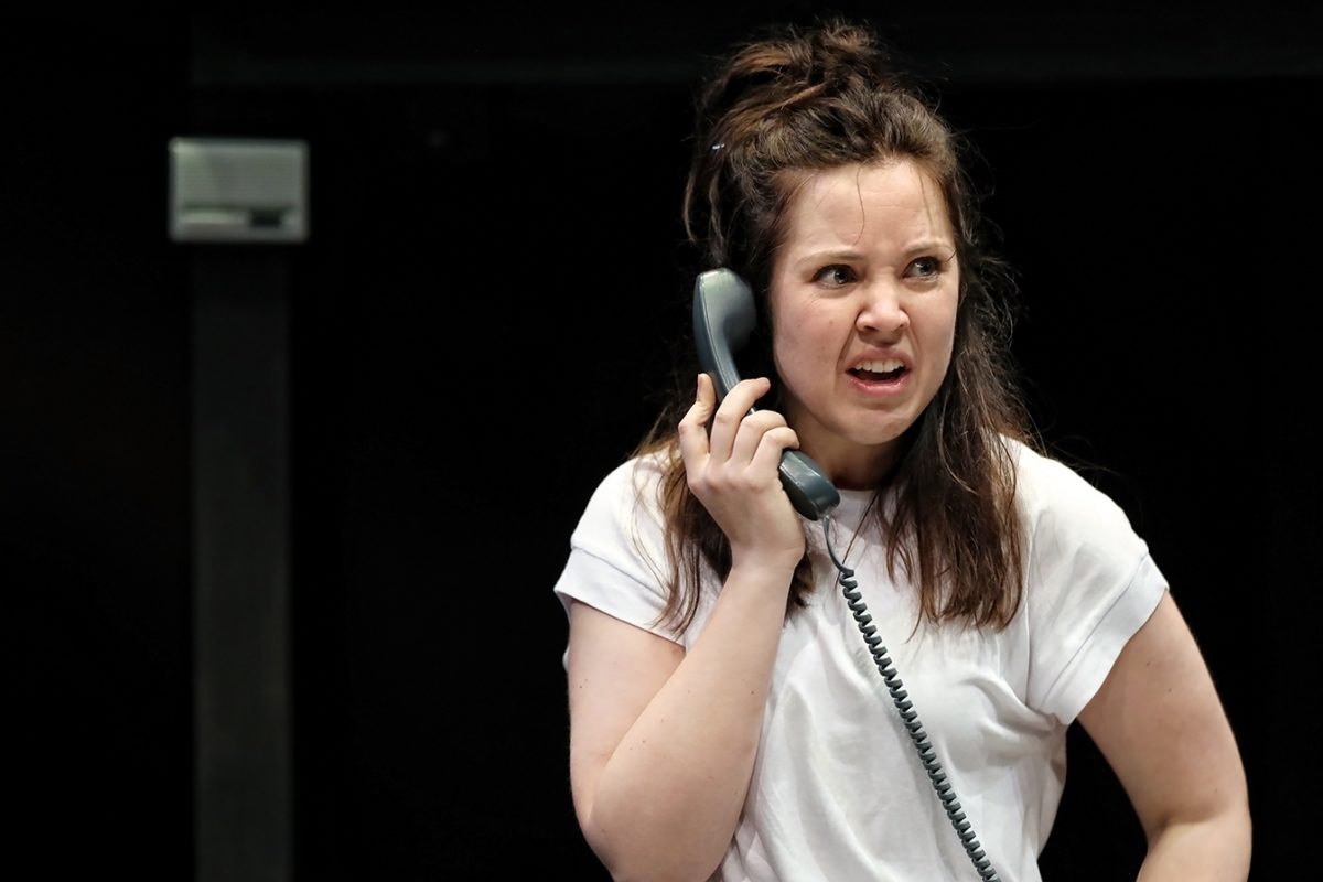 A woman with long brown hair in a white T-shirt holds a corded telephone to her ear, looking upset or frustrated, with her brow furrowed and lips parted, against a dark background.