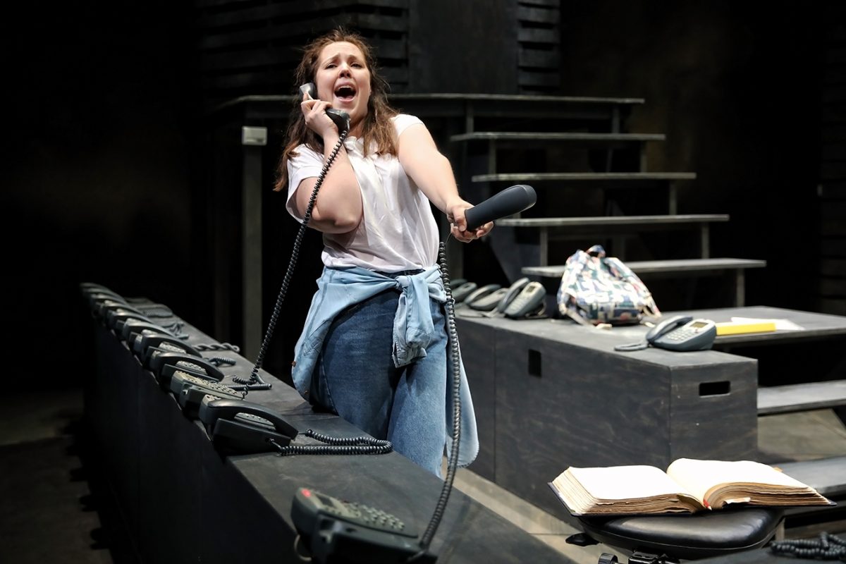 A woman stands on stage holding two mobiles, appearing distressed as she speaks. She wears jeans with a shirt tied around her waist. Multiple mobiles are on the desk, and an open book and rucksack are nearby.