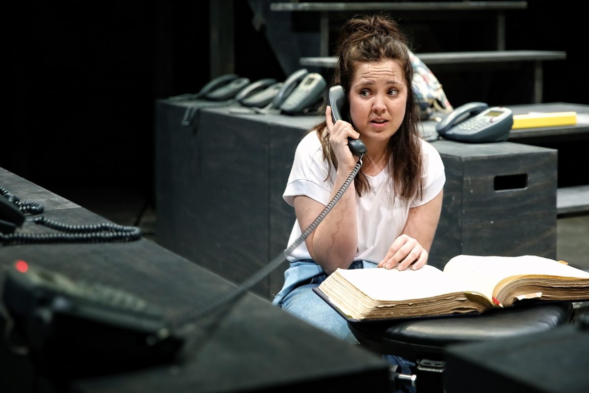 A woman in a white shirt sits at a table, holding a corded telephone to her ear and looking concerned. She has an open book in front of her, and several other telephones are on nearby tables.