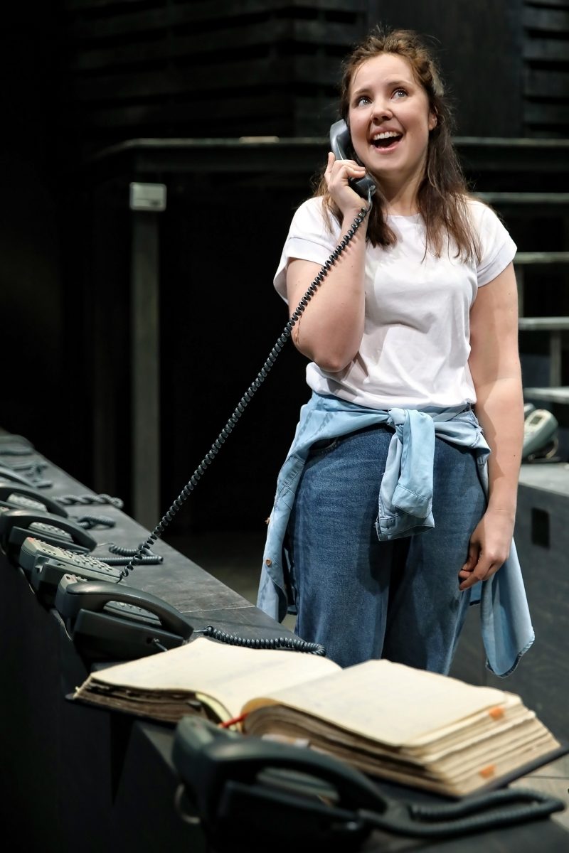 A young woman in a white T-shirt and jeans stands on stage, smiling as she talks on a corded telephone. Open notebooks and several telephones are on the table in front of her, with a dark theatre set in the background.
