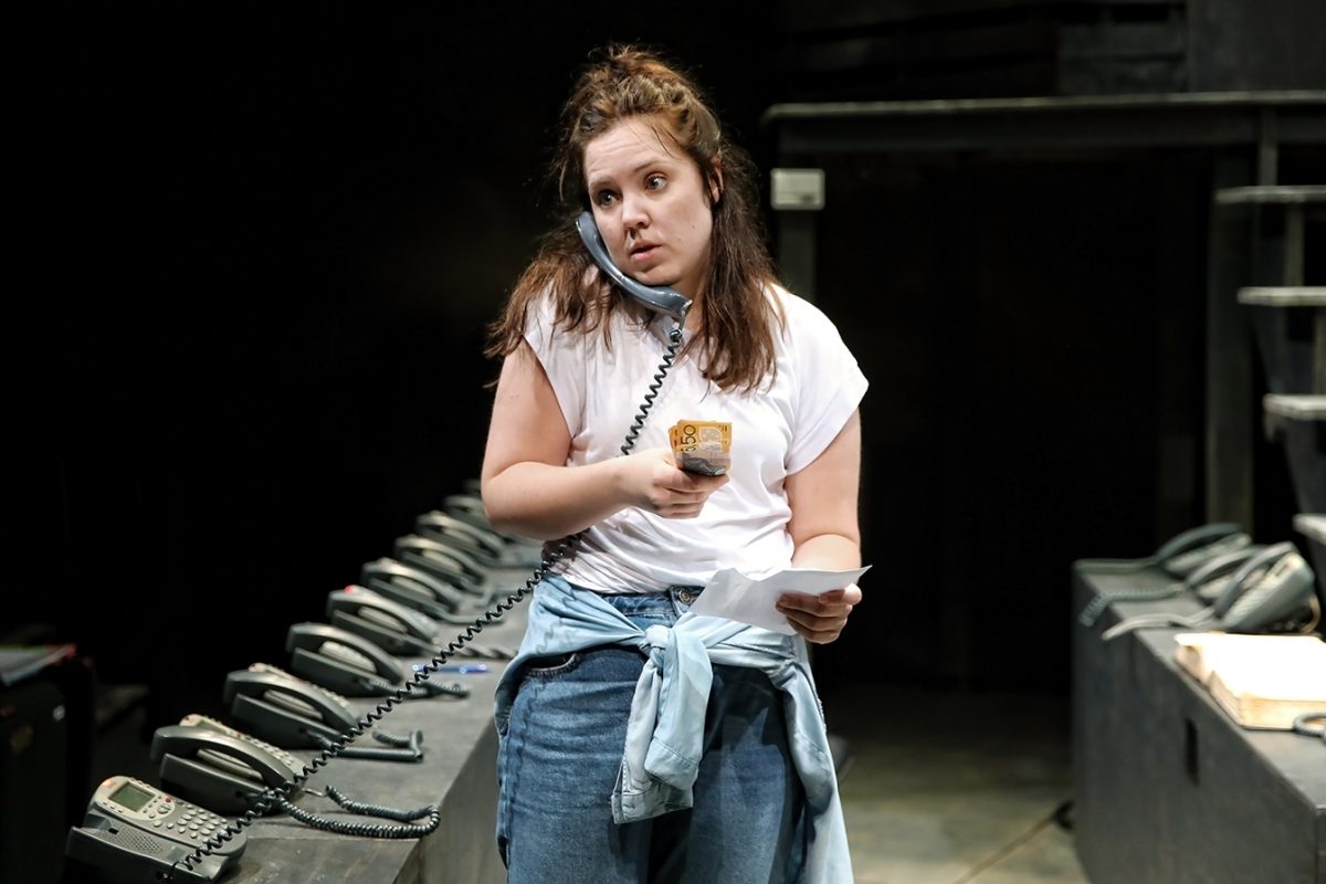 A woman stands in a dimly lit room holding papers, talking on a corded phone with the receiver wedged between her shoulder and ear. She wears jeans, a white T-shirt, and a denim shirt tied at her waist. Multiple telephones line the table beside her.