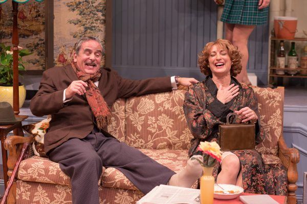 An older man and woman sit laughing together on a floral sofa in a cosy, vintage-style sitting room. The man wears a suit and scarf, and the woman holds her hand to her chest, both appearing joyful and relaxed.