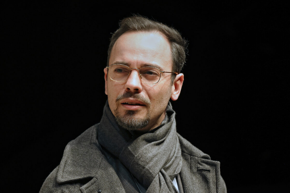 A man with glasses, short dark hair, and a goatee, wearing a grey scarf and a herringbone coat, looks slightly to the side against a black background.