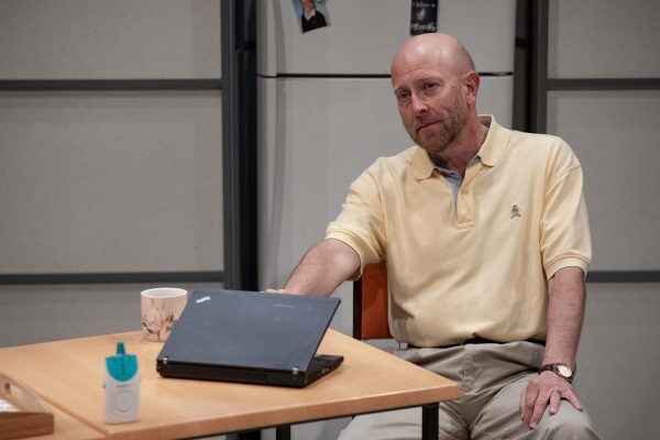 A man with a bald head and beard, wearing a light yellow polo shirt and beige trousers, sits at a table with a closed laptop, a floral mug, and a small device. A fridge is visible in the background.