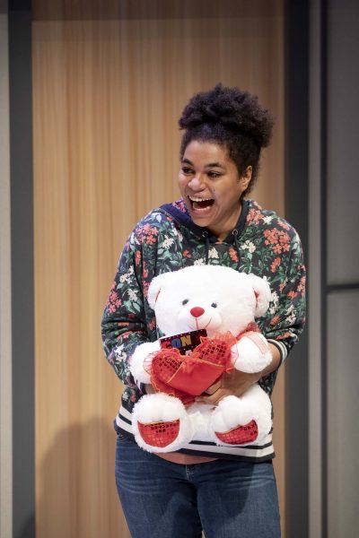 A smiling woman wearing a floral hoodie holds a white teddy bear with red accents and a heart that says I ♥ U. She stands in front of a neutral background, looking happy and joyful.