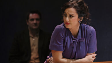 A woman in a purple blouse leans on a table, looking to the side with a concerned expression. A man, blurred and sitting in the background, watches her. The scene appears tense and dramatic.