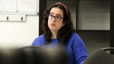 A woman with long dark hair, glasses, and a patterned headband, wearing a blue jumper, sits at a table and looks attentively to the side in a classroom or office setting.