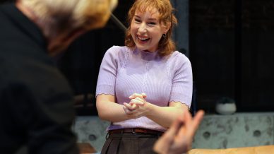 A woman with red hair wearing a lavender jumper smiles brightly with her hands clasped, facing a man with blond hair whose back is to the camera. They appear to be having an animated conversation indoors.