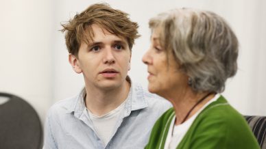 A young man with tousled hair looks attentively at an older woman with short grey hair, who is wearing a green cardigan and speaking, both seated indoors.