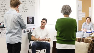 A man sits on a chair talking and gesturing with his hands whilst three other people stand or sit around him in a casual room with whiteboards and notes on the walls.