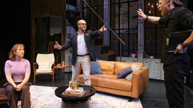 Three people are on a stage set designed like a modern living room. A seated woman looks forward, a man in glasses stands pointing, and another man in a cap stands with a hand raised, holding a notebook.