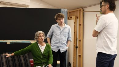 Three people are in conversation indoors. An older woman in a green jumper is seated, while two men stand listening, one gesturing with his hand. The setting appears to be a workshop or rehearsal space.