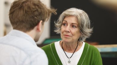 An older woman with grey hair and a green cardigan looks at a younger person with concern while listening, in an indoor setting. The woman appears engaged and empathetic during their conversation.