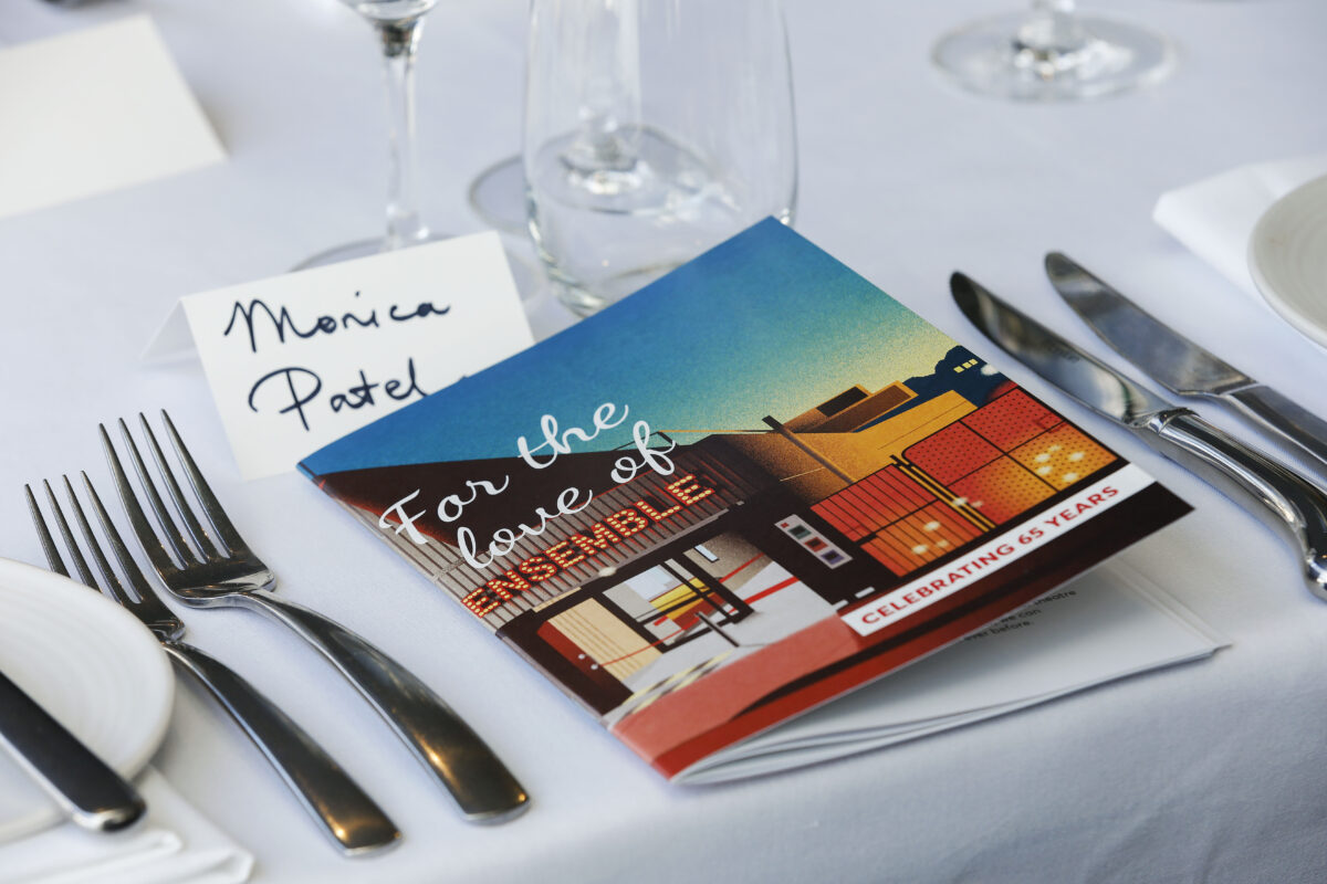 A place setting with a booklet titled For the love of MUSEUM, a name card reading Monica Patel, wine glasses, cutlery, and plates on a white tablecloth. The booklet notes Celebrating 65 years.