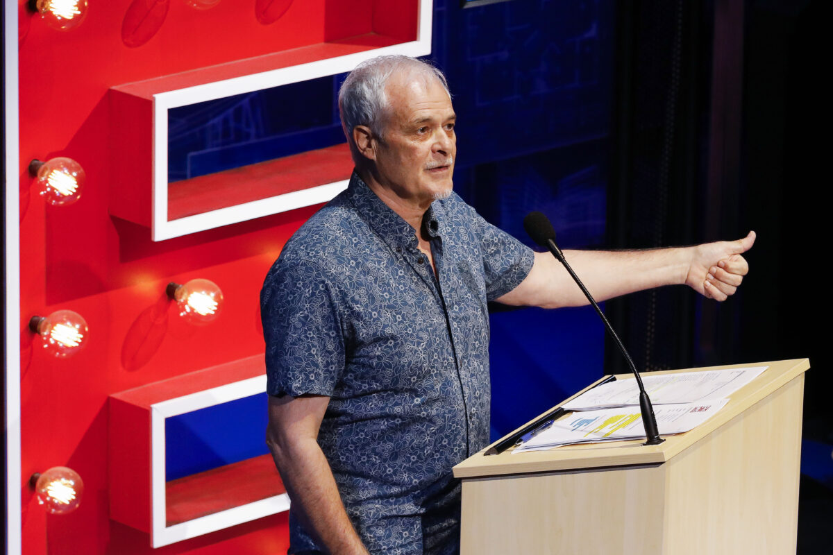 An older man with grey hair speaks at a lectern, gesturing with his right hand. Behind him is a large, illuminated red letter E. Papers are spread out on the lectern.