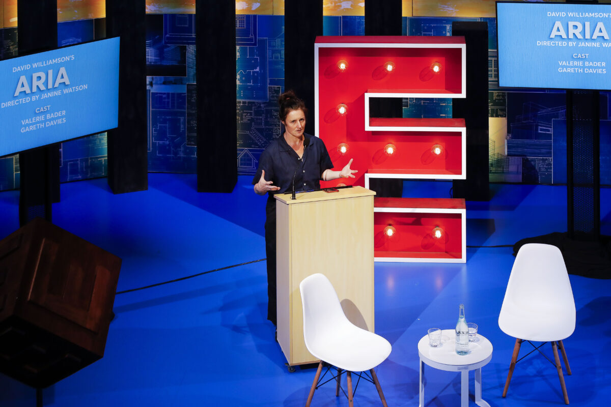 A woman speaks at a lectern on stage, gesturing with her hands. Behind her is a large red letter E lit with bulbs, blue screens with text, and two empty white chairs with a small round table and a water bottle.