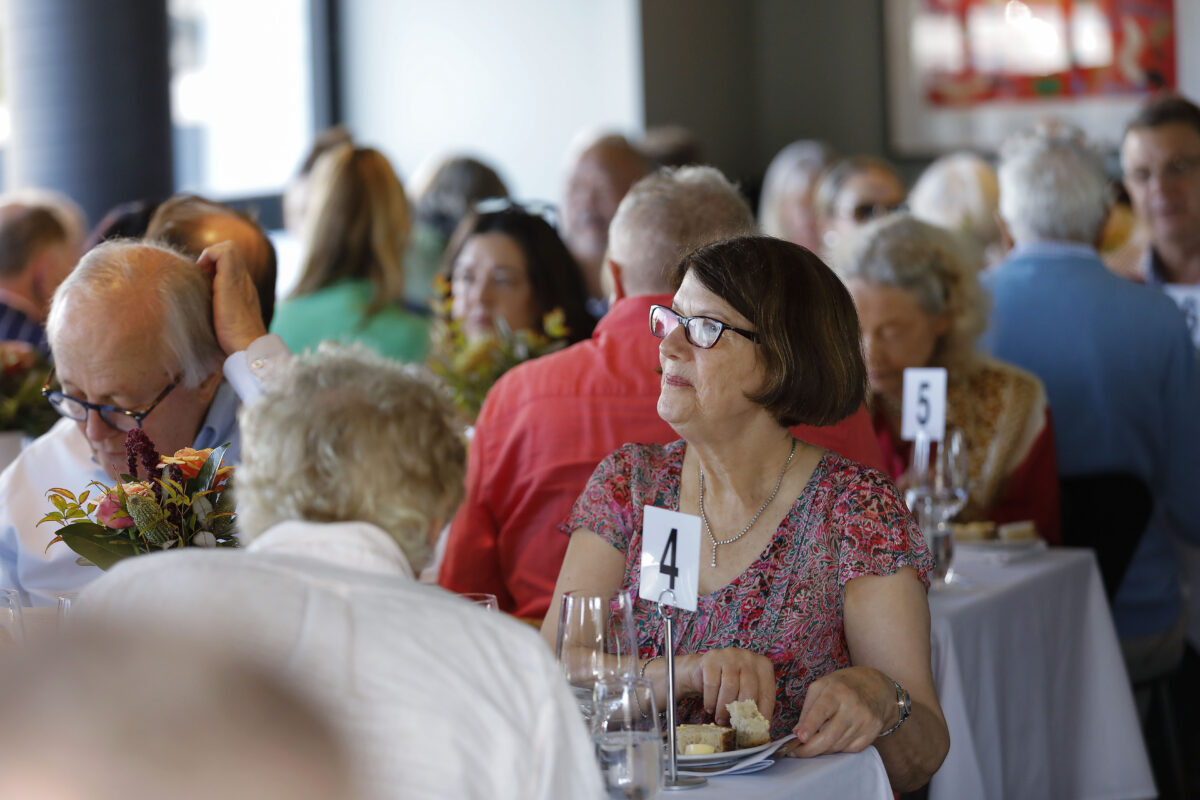 A group of people are seated at tables covered with white tablecloths, dining together in a bright, busy restaurant. A woman in glasses and a floral top sits at table number 4, looking ahead thoughtfully.