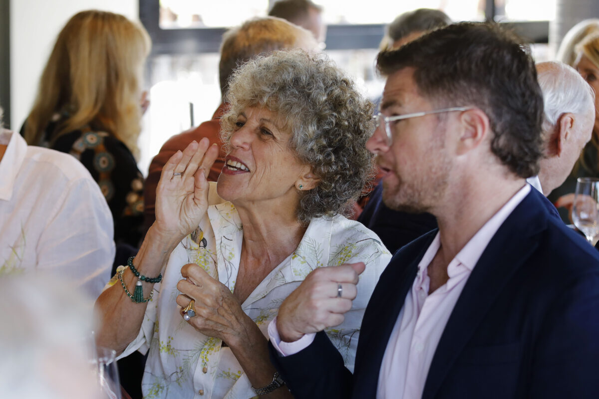 A woman with curly grey hair smiles and gestures while talking to a man in glasses and a navy blazer at a lively indoor gathering. Other people are sitting and conversing in the background.