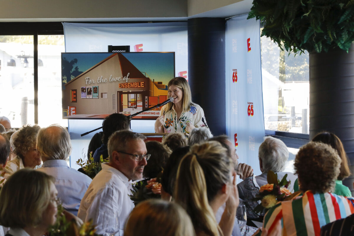 A woman speaks at a lectern to a seated audience in a brightly lit room; behind her is a large screen displaying the words For the love of ENSEMBLE and an image of a building.