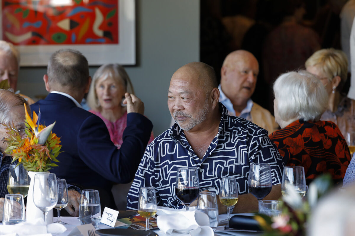 A man in a patterned shirt sits at a table with wine glasses and flowers, surrounded by people engaged in conversation at a social gathering in a well-lit room.