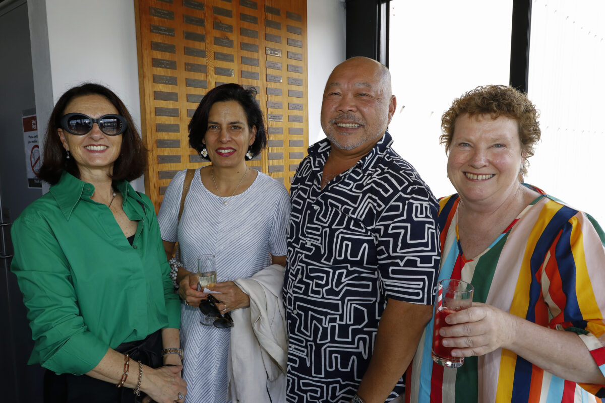 Four adults stand closely together, smiling at the camera. One woman wears a green blouse and sunglasses, another a light striped dress, a man wears a patterned shirt, and a woman in a colourful striped top holds a drink.