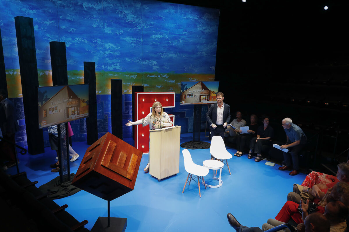 A woman speaks at a lectern on a blue stage with house illustrations and props, while a man stands nearby. An audience sits to the right, watching the presentation. The background features a colourful painted wall.