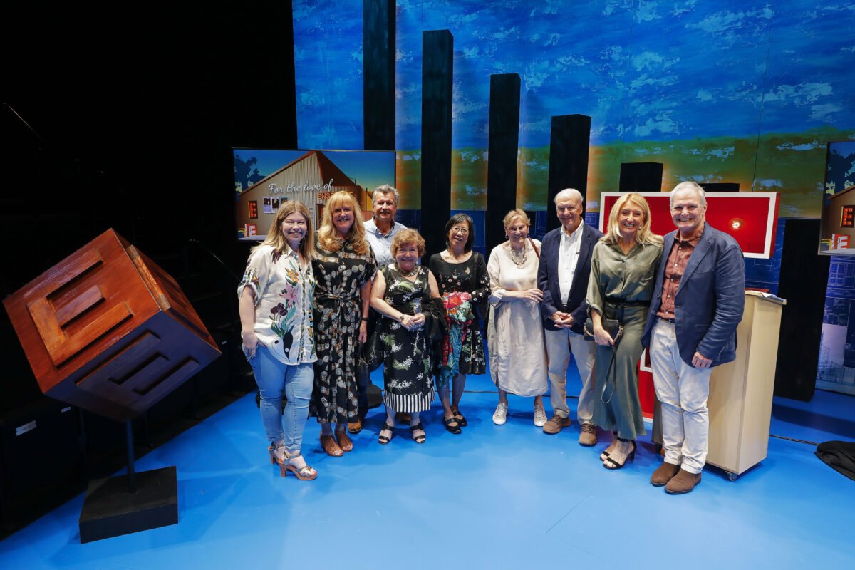 A group of ten adults stand together and smile on a brightly lit stage with blue flooring and a painted sky backdrop. There are tall black columns and a large geometric wooden sculpture beside them.