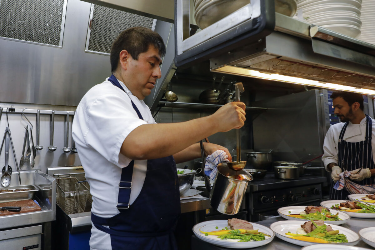 A chef in a white uniform pours sauce onto plated dishes in a commercial kitchen, whilst another chef works in the background. Plates of food with meat and vegetables are lined up on the counter.