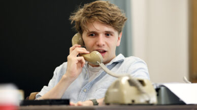 A young man with tousled hair talks on a beige rotary phone at a desk, looking focused. Papers and another phone are on the desk, and there is a blurred background with a blackboard and curtain.