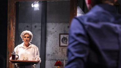 An older woman stands holding a tray with a teapot and cups, facing a person with short hair in the foreground. They are in a dimly lit room with a black sofa and wooden table.