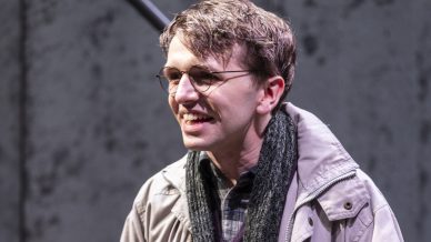 A young man wearing glasses, a light grey coat, scarf, and carrying a brown leather bag smiles whilst standing indoors in front of a concrete wall and staircase.