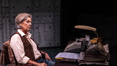 An older person with grey hair sits at a cluttered desk with a typewriter, telephone, and papers, looking thoughtful. The background is a concrete wall, and the lighting is dramatic.
