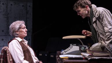 An older person sits at a desk with a typewriter, looking up at a younger person standing and leaning forward, speaking intensely. The setting appears to be a dramatic stage play.