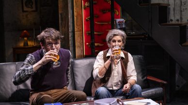 Two people sit on a dark settee in a cosy, dimly lit room, each holding a glass of beer. Behind them is a cupboard displaying various knives, and a table in front of them holds jars, papers, and a gift box.