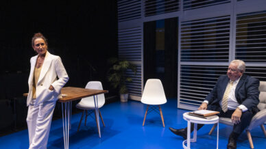 A woman in a white suit stands by a desk, speaking, while an older man in a suit sits on a chair next to a small table. The scene takes place on a blue floor, with modern furniture and window blinds in the background.