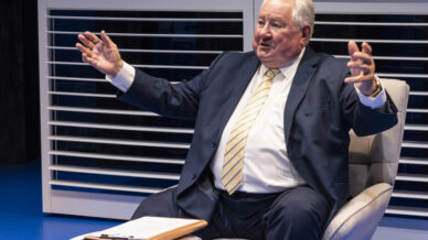 An older man in a dark suit and yellow striped tie sits in a modern grey armchair with his arms outstretched, smiling. A small white table with a folder and pen is in front of him, with white blinds in the background.