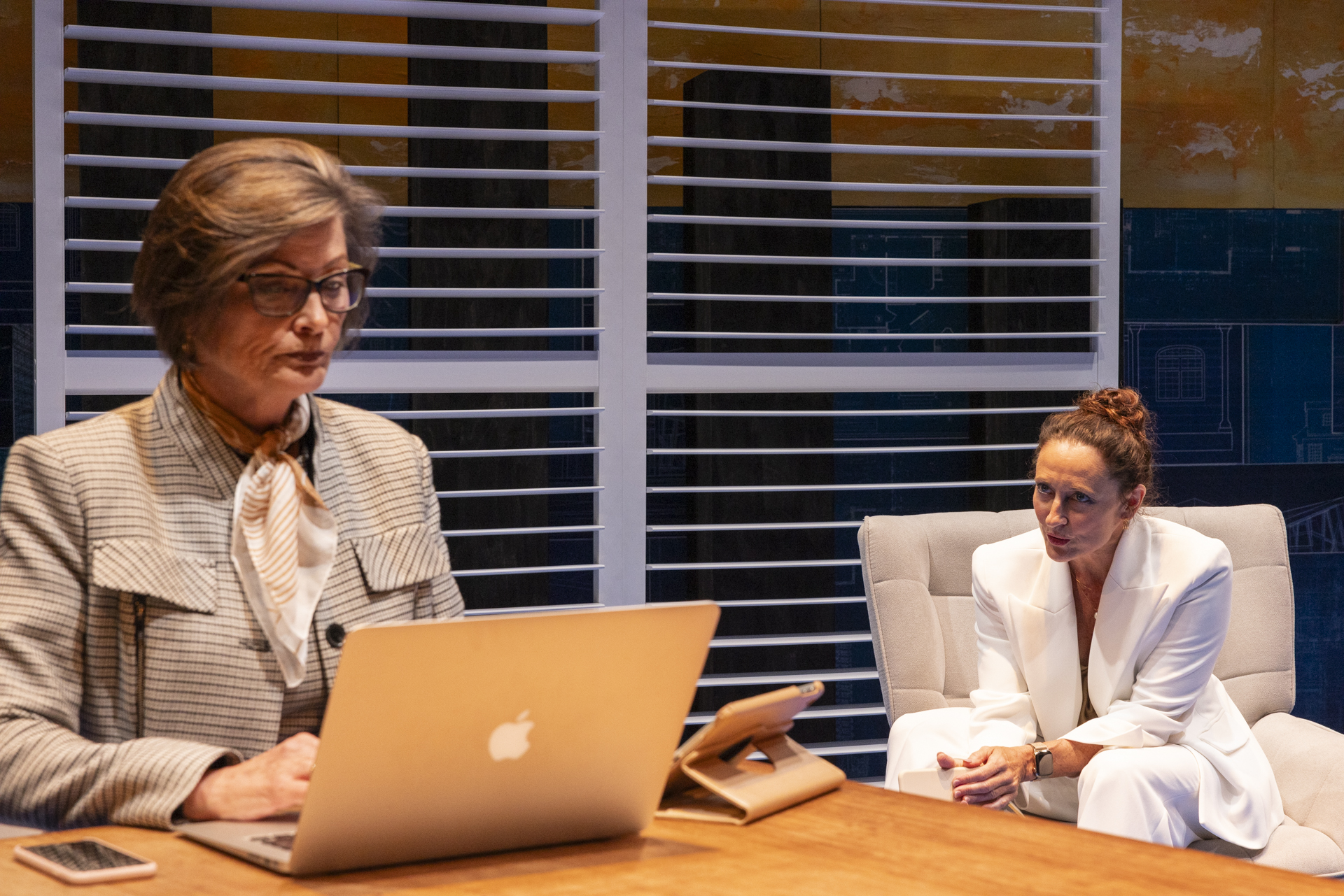Two women in business attire sit at a desk. One, with short hair and glasses, works on a laptop. The other, with curly hair in a bun, sits opposite her, leaning forward and listening intently. Blinds and cityscape in the background.