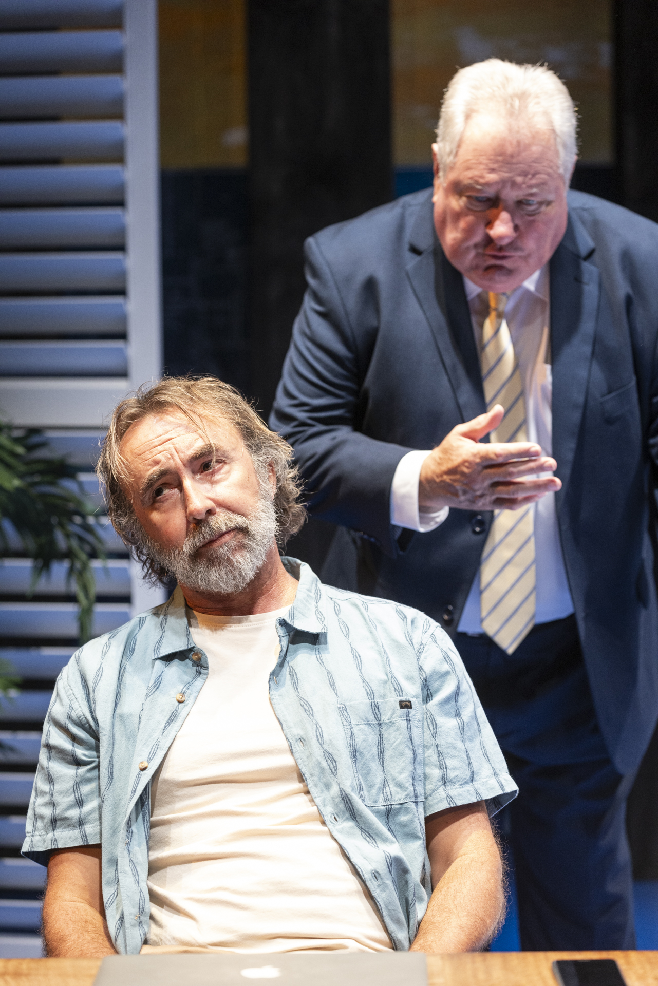 A man in a casual shirt sits at a table looking concerned while another man in a suit stands behind him, gesturing emphatically as if speaking or arguing.