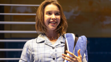 A woman with shoulder-length brown hair, wearing a light blue checked shirt, smiles whilst holding striped towels. She stands in front of a window with horizontal blinds and a warmly lit background.