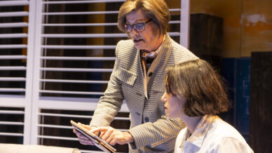 Two women indoors: one standing and pointing at a tablet, the other seated and looking at the screen. The background features window blinds and warm lighting.