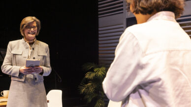A woman in a grey suit smiles and holds a tablet while speaking to another person in a white shirt. They are indoors with modern decor, potted plants, and chairs visible in the background.