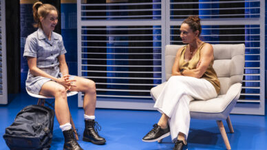 A teenage girl in a school uniform and boots sits on a bench, smiling, with a rucksack at her feet. An older woman in a tan top and white trousers sits on a chair nearby, looking at her with a serious expression.